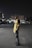 A person stands outdoors at night on an empty street, wearing a yellow sports jersey over a hoodie. The background consists of dimly lit buildings and streetlights, with a car parked on one side.