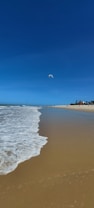 A serene beachfront with gentle ocean waves caressing the golden sand under a clear blue sky. Several kites can be seen in the distance, suggesting kite surfing activity. People are visible along the shoreline, enjoying the sunny day.