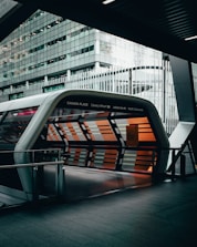 A modern architectural structure with a tunnel-like entrance covered in glass panels. The panels display a pattern of orange and gray stripes, giving a sleek, futuristic appearance. The surrounding building is made of glass and steel, featuring reflective surfaces. The space appears urban, likely part of a transportation hub.