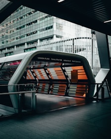 A modern architectural structure with a tunnel-like entrance covered in glass panels. The panels display a pattern of orange and gray stripes, giving a sleek, futuristic appearance. The surrounding building is made of glass and steel, featuring reflective surfaces. The space appears urban, likely part of a transportation hub.
