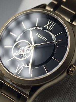 A close-up view of a Fossil brand wristwatch showcasing a detailed mechanical design with visible gears through the transparent watch face. The watch features Roman numerals and sleek metallic hands, set against a dark background, all encased in a metallic frame with a matching metal strap.