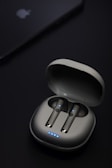 A pair of wireless earbuds are seated in an open charging case. The earbuds and case have a sleek, modern design with small blue LED lights indicating they are powered or charging. Nearby, a device that resembles a smartphone or tablet is partially visible with a logo on it.
