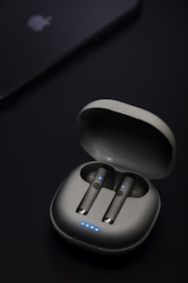 A pair of wireless earbuds are seated in an open charging case. The earbuds and case have a sleek, modern design with small blue LED lights indicating they are powered or charging. Nearby, a device that resembles a smartphone or tablet is partially visible with a logo on it.