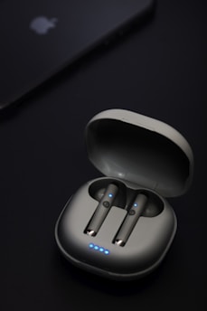 A pair of wireless earbuds are seated in an open charging case. The earbuds and case have a sleek, modern design with small blue LED lights indicating they are powered or charging. Nearby, a device that resembles a smartphone or tablet is partially visible with a logo on it.