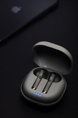 A pair of wireless earbuds are seated in an open charging case. The earbuds and case have a sleek, modern design with small blue LED lights indicating they are powered or charging. Nearby, a device that resembles a smartphone or tablet is partially visible with a logo on it.