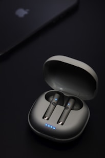 A pair of wireless earbuds are seated in an open charging case. The earbuds and case have a sleek, modern design with small blue LED lights indicating they are powered or charging. Nearby, a device that resembles a smartphone or tablet is partially visible with a logo on it.