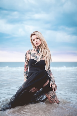A blonde woman with extensive tattoos is kneeling in shallow water near a beach. She is wearing a black dress, and the background features the ocean with waves and a cloudy sky.