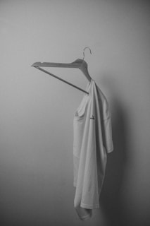 A white t-shirt is hanging on a plain wooden hanger against a smooth, light-colored wall. The lighting is soft, creating gentle shadows and giving the scene a minimalist and serene appearance.