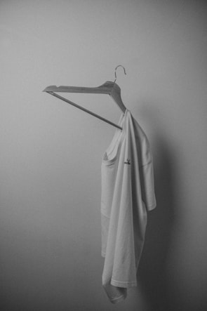 A white t-shirt is hanging on a plain wooden hanger against a smooth, light-colored wall. The lighting is soft, creating gentle shadows and giving the scene a minimalist and serene appearance.