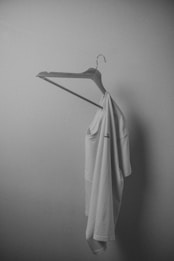 A white t-shirt is hanging on a plain wooden hanger against a smooth, light-colored wall. The lighting is soft, creating gentle shadows and giving the scene a minimalist and serene appearance.