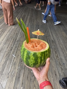 A hand holding a hollowed-out watermelon filled with a blended drink adorned with a small paper cocktail umbrella and two tall leaves. The setting appears to be a wooden boardwalk with people walking in the background, dressed casually in summer attire.