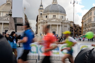 A busy city square with several blurred motion figures running in the foreground, suggesting a race or marathon. A photographer in the background captures the event. The scene is set against historic European architecture with a large dome and intricate details, indicating an iconic urban location. A crowd of spectators lines the path.