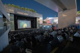 A large outdoor theater is filled with people seated on tiered steps, watching a projected image on a big screen. The setting is urban, with modern architecture and lit signage visible. Trees are present along the upper edges, and the sky is clear.