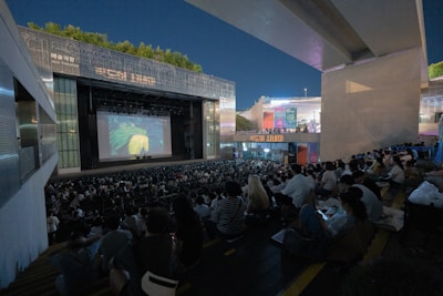 A large outdoor theater is filled with people seated on tiered steps, watching a projected image on a big screen. The setting is urban, with modern architecture and lit signage visible. Trees are present along the upper edges, and the sky is clear.