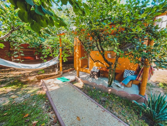 A cozy outdoor setting with a hammock hanging between trees and a small, rustic wooden cabin nestled in lush greenery. A gravel path leads to the cabin, and a person relaxes on a hammock with vibrant pillows adding a touch of color. The area is surrounded by trees and plants, creating a peaceful and inviting atmosphere.