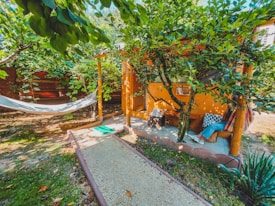 A cozy outdoor setting with a hammock hanging between trees and a small, rustic wooden cabin nestled in lush greenery. A gravel path leads to the cabin, and a person relaxes on a hammock with vibrant pillows adding a touch of color. The area is surrounded by trees and plants, creating a peaceful and inviting atmosphere.