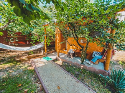 A cozy outdoor setting with a hammock hanging between trees and a small, rustic wooden cabin nestled in lush greenery. A gravel path leads to the cabin, and a person relaxes on a hammock with vibrant pillows adding a touch of color. The area is surrounded by trees and plants, creating a peaceful and inviting atmosphere.