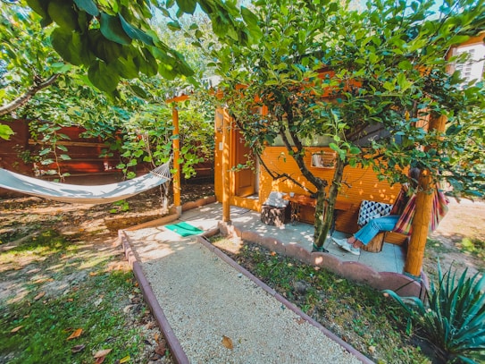 A cozy outdoor setting with a hammock hanging between trees and a small, rustic wooden cabin nestled in lush greenery. A gravel path leads to the cabin, and a person relaxes on a hammock with vibrant pillows adding a touch of color. The area is surrounded by trees and plants, creating a peaceful and inviting atmosphere.