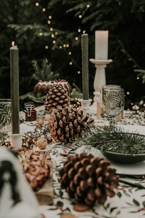 A festive table setting featuring large pinecones, green candles, and a decorative cake with pine branches and berries. Glass goblets with a leafy pattern are placed alongside a white candle on a stand. The background includes small decorative lights, giving a cozy, warm atmosphere.
