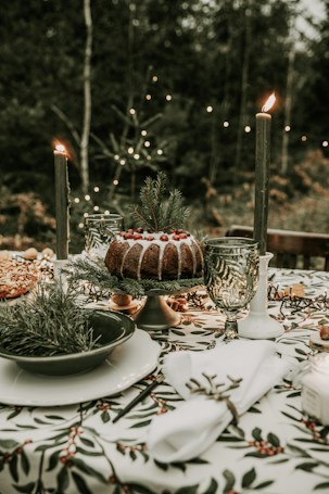 A festive table setting arranged outdoors with a central cake topped with cranberries and greenery. Two lit candles, decorative glassware, and a patterned tablecloth featuring holly leaves and berries adorn the table. Pine branches add to the winter holiday theme, with fairy lights softly illuminating the background.