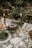 A festive dining table arrangement is set outdoors with a holiday theme. The table is adorned with a patterned tablecloth featuring greenery and red berries. A bundt cake decorated with white icing and pomegranate seeds sits as a centerpiece with sprigs of evergreen. Two lit candles in white holders add warmth, while crystal glassware and neatly arranged tableware complete the setting. In the background, a small decorated tree with fairy lights enhances the festive atmosphere.