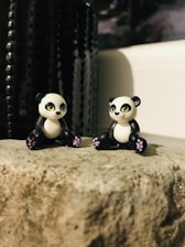 Two small panda figurines with cute expressions are placed on a textured rock surface. The pandas have black and white coloring with pink accents on their paws. A dark, textured background creates a contrast with the lighter foreground.