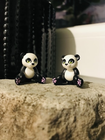 Two small panda figurines with cute expressions are placed on a textured rock surface. The pandas have black and white coloring with pink accents on their paws. A dark, textured background creates a contrast with the lighter foreground.