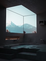 A modern, minimalist bathroom features a sunken bathtub with a mountain view through a large floor-to-ceiling window. The space is adorned with several candles, and towels are neatly stacked on a shelf. A stool is positioned nearby, and the overall ambiance is serene and calming.