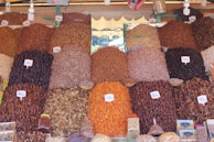 A vibrant market stall displays a variety of dried fruits, nuts, and spices organized in neat piles. Each pile is labeled with a price tag in the local currency. The arrangement creates a visually appealing pattern with various textures and colors.