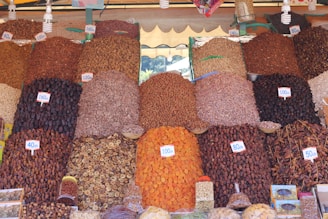 A vibrant market stall displays a variety of dried fruits, nuts, and spices organized in neat piles. Each pile is labeled with a price tag in the local currency. The arrangement creates a visually appealing pattern with various textures and colors.