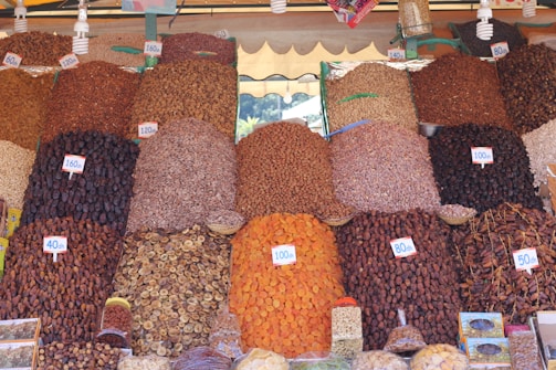 A vibrant market stall displays a variety of dried fruits, nuts, and spices organized in neat piles. Each pile is labeled with a price tag in the local currency. The arrangement creates a visually appealing pattern with various textures and colors.