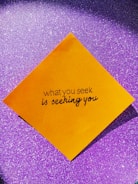A yellow square paper with a motivational quote written in black script font is placed against a sparkly purple background.