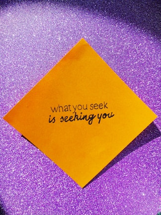 A yellow square paper with a motivational quote written in black script font is placed against a sparkly purple background.