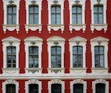 A facade of a historic building featuring six large, arched windows with intricate white stucco detailing set against a deep red background. The windows are symmetrically arranged in two rows with decorative elements above and below each window. The architectural style suggests elegance and classical influences.