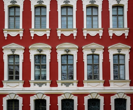 A facade of a historic building featuring six large, arched windows with intricate white stucco detailing set against a deep red background. The windows are symmetrically arranged in two rows with decorative elements above and below each window. The architectural style suggests elegance and classical influences.