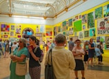 A vibrant art gallery room with high ceilings, ornate moldings, and brightly colored walls displaying an eclectic mix of artwork. People are engaged in discussions, and some are examining the artworks closely. The atmosphere suggests active interest and engagement with the art pieces.