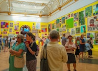 A vibrant art gallery room with high ceilings, ornate moldings, and brightly colored walls displaying an eclectic mix of artwork. People are engaged in discussions, and some are examining the artworks closely. The atmosphere suggests active interest and engagement with the art pieces.