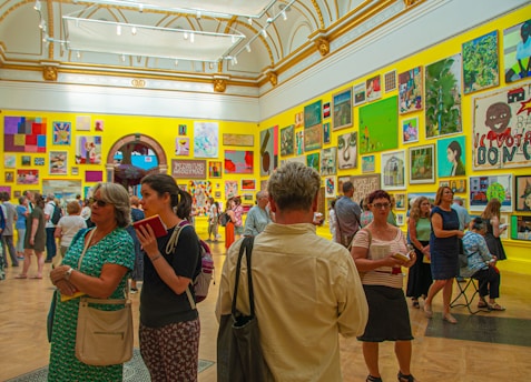 A vibrant art gallery room with high ceilings, ornate moldings, and brightly colored walls displaying an eclectic mix of artwork. People are engaged in discussions, and some are examining the artworks closely. The atmosphere suggests active interest and engagement with the art pieces.