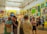A vibrant art gallery room with high ceilings, ornate moldings, and brightly colored walls displaying an eclectic mix of artwork. People are engaged in discussions, and some are examining the artworks closely. The atmosphere suggests active interest and engagement with the art pieces.