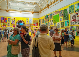 A vibrant art gallery room with high ceilings, ornate moldings, and brightly colored walls displaying an eclectic mix of artwork. People are engaged in discussions, and some are examining the artworks closely. The atmosphere suggests active interest and engagement with the art pieces.