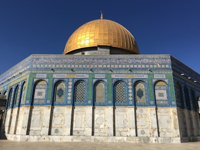 A magnificent structure featuring a large golden dome sitting atop an intricately decorated building. The walls are adorned with detailed blue and green geometric patterns and calligraphy, indicative of Islamic architecture.