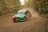 A rally car speeds along a dirt track surrounded by dense forest. The car is kicking up a cloud of dust and features vibrant colors with sponsor logos. The setting suggests an off-road race in an autumnal environment.