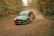 A rally car speeds along a dirt track surrounded by dense forest. The car is kicking up a cloud of dust and features vibrant colors with sponsor logos. The setting suggests an off-road race in an autumnal environment.