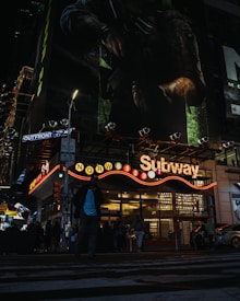 A nighttime urban scene featuring a subway entrance illuminated by multicolored lights. A large video game advertisement is prominently displayed on a building above. Pedestrians are seen walking across a zebra crossing in the foreground.