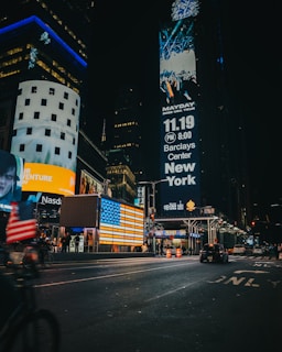 A bustling city scene at night with large digital billboards displaying advertisements. The streets are well-lit with neon lights reflecting off the wet pavement. People are visible walking and cars are driving down the street. A prominent American flag display is illuminated beside the billboards.