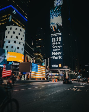 A bustling city scene at night with large digital billboards displaying advertisements. The streets are well-lit with neon lights reflecting off the wet pavement. People are visible walking and cars are driving down the street. A prominent American flag display is illuminated beside the billboards.