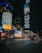 A bustling city scene at night with large digital billboards displaying advertisements. The streets are well-lit with neon lights reflecting off the wet pavement. People are visible walking and cars are driving down the street. A prominent American flag display is illuminated beside the billboards.