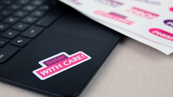 A laptop keyboard with a sticker saying 'SHARE WITH CARE!' attached to the surface. Beside the laptop, there is a sheet of paper with more similar stickers visible.