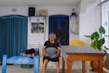 An elderly woman sits smiling in a shaded, outdoor area. She is on a yellow plastic chair beside a wooden table covered with a gray patterned cloth. The setting includes blue and white elements, with a vibrant blue curtain, blue window grilles, and white walls. There are various decorative items on the walls, including a painting, a butterfly ornament, and a walking stick. Potted plants add greenery to the scene, and a blue chair is visible in the foreground.