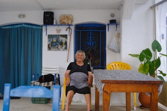 An elderly woman sits smiling in a shaded, outdoor area. She is on a yellow plastic chair beside a wooden table covered with a gray patterned cloth. The setting includes blue and white elements, with a vibrant blue curtain, blue window grilles, and white walls. There are various decorative items on the walls, including a painting, a butterfly ornament, and a walking stick. Potted plants add greenery to the scene, and a blue chair is visible in the foreground.